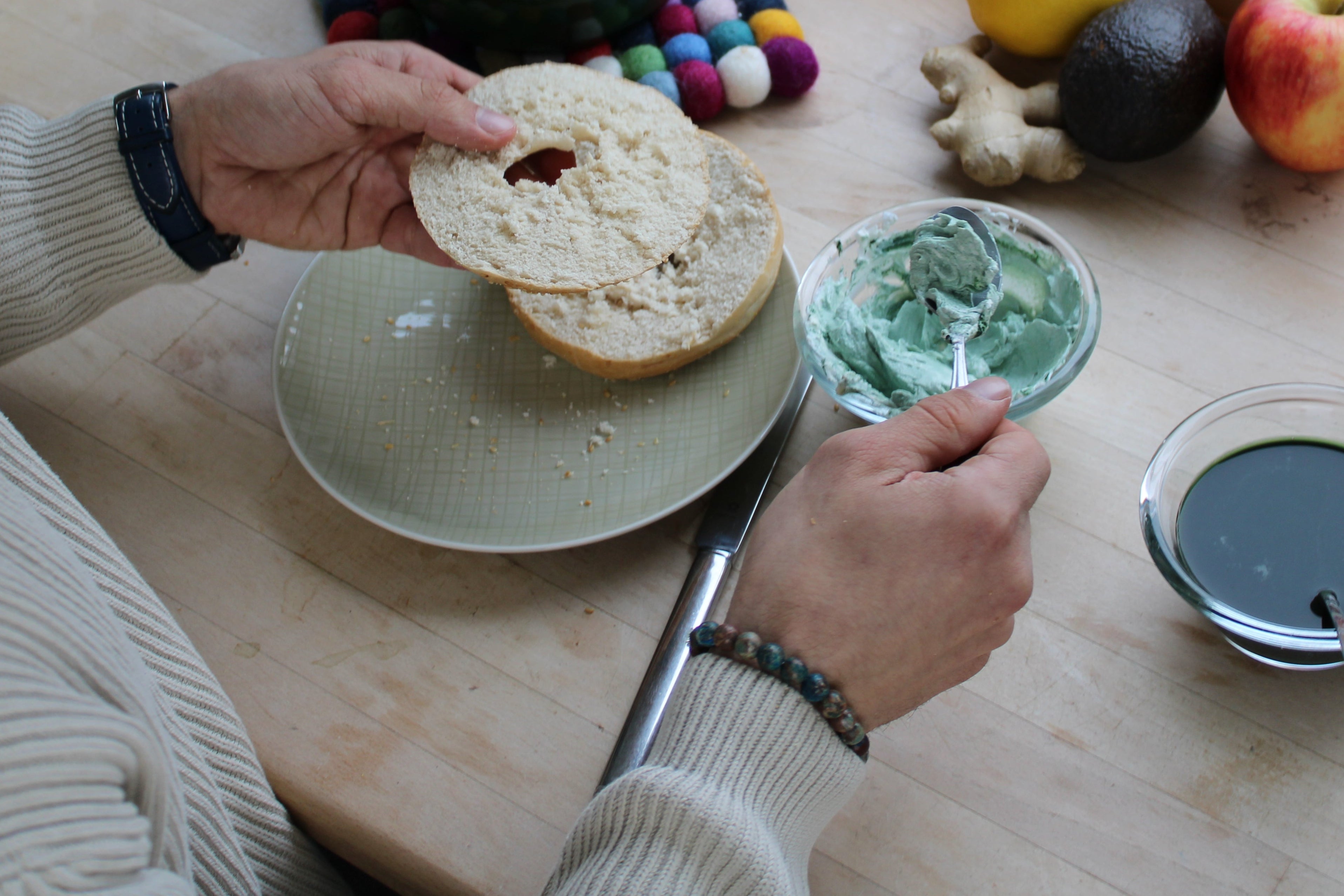 Person, die gerade frischkäse auf einen Bagel schmiert: Der Frischkäse hat eine grüne Farbe durch die Mischung mit Spirulina, im Hintergrund eine Spirulina-Farm auf einem Fensterbrett.