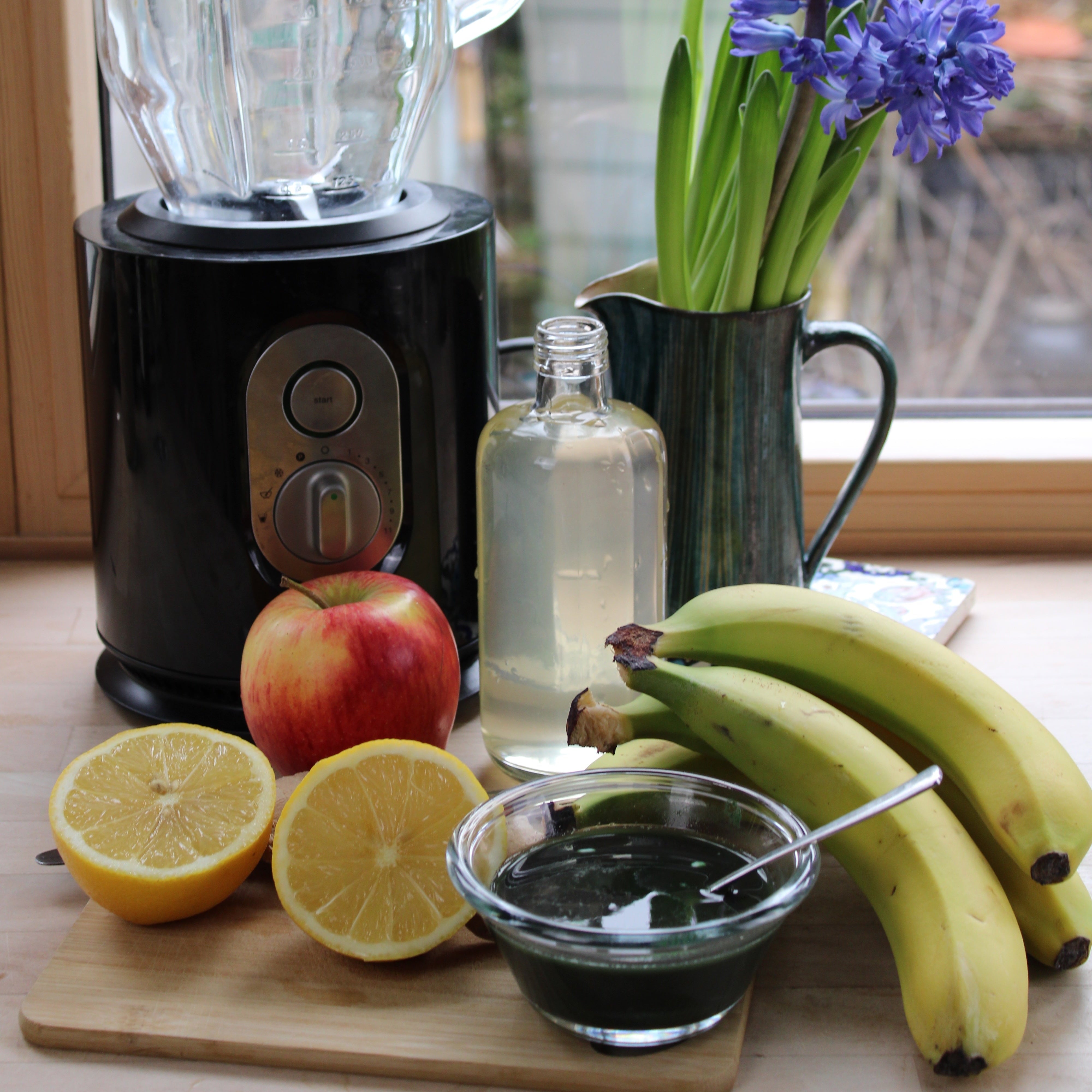 Frisches Obst (Zitrone, Banane und Apfel) mit einer Schüssel Spirulina im Vordergrund: Im Hintergrund ein Mixer, eine Flasche und eine Blume, die eine frische, gesunde Atmosphäre vermitteln.