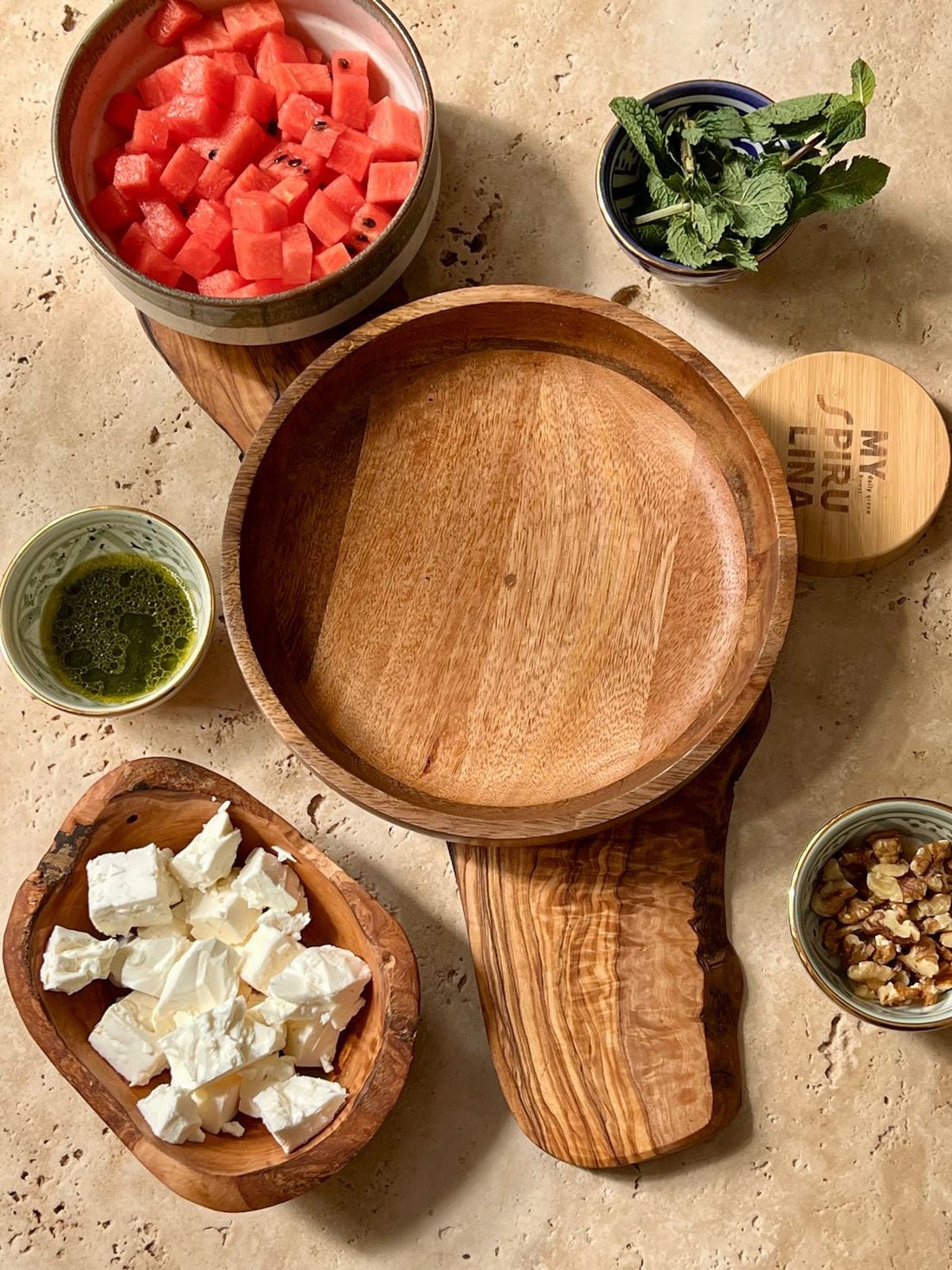 Wassermelonenwürfel, Feta, Walnüsse, Minze und ein Schälchen grünes Spirulina Dressing in Holzschalen auf hellem Stein