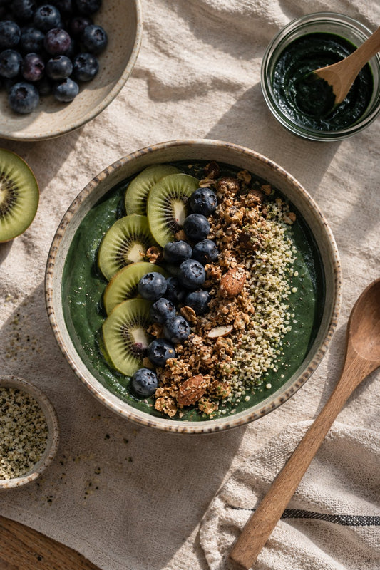 Grüne Smoothie Bowl mit frischer Spirulina von oben, belegt mit Kiwischeiben, Blaubeeren, Granola und Hanfsamen auf einer Leinentischdecke